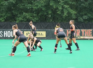 Several athletes in matching sports uniforms are on a bright green field, engaged in a field hockey game. They appear focused and are holding hockey sticks, with some bending slightly as they prepare to play. Trees and a billboard are visible in the background.