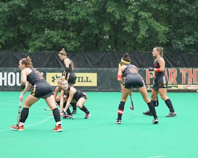 Several athletes in matching sports uniforms are on a bright green field, engaged in a field hockey game. They appear focused and are holding hockey sticks, with some bending slightly as they prepare to play. Trees and a billboard are visible in the background.