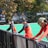 A group of athletes wearing orange uniforms are on the sideline of a sports field, resting against a fence. Behind them, a large digital scoreboard displays team names and scores. The field is bright green, and several banners with sponsor logos and championship wins are visible. The athletes appear to be taking a break, possibly before or during a field hockey game.