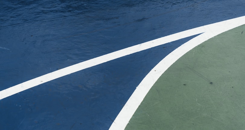A close-up view of a sports court featuring distinctly marked lines, with a dark blue surface contrasted by a curve of green. The white lines delineate the boundary between the two areas, creating an abstract visual effect.