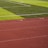 A sports field with vivid green grass divided into sections by white lines in the background. In the foreground, a track with a reddish-brown surface is marked with numbers and lines.