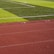A sports field with vivid green grass divided into sections by white lines in the background. In the foreground, a track with a reddish-brown surface is marked with numbers and lines.