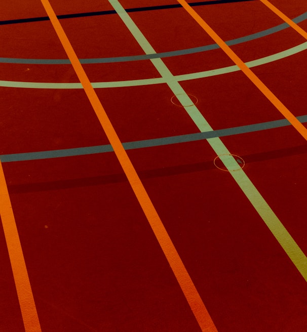 A sports court with intersecting lines in orange and blue, creating geometric patterns on a reddish-brown surface.