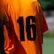 A person is wearing an orange sports jersey with the number 16 printed in black on the back. The background is blurred, but it appears to be an outdoor environment, possibly a sports field.
