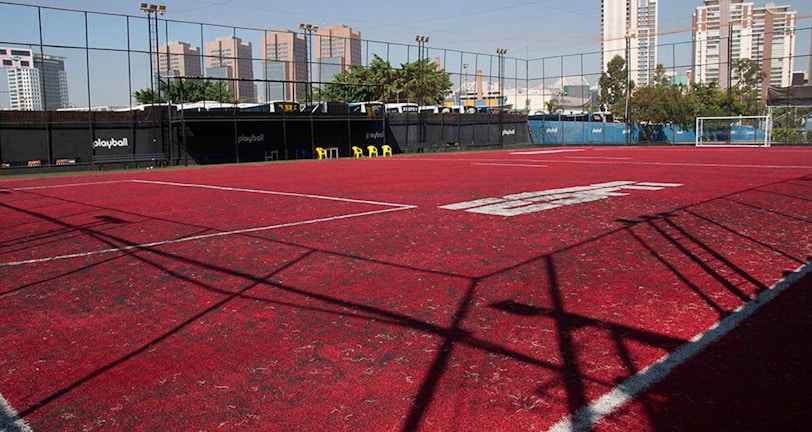 A sports court with a red playing surface and white lines, surrounded by a black mesh fence. In the background, tall buildings are visible, along with some trees and a few yellow chairs positioned near the fence.